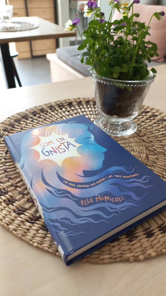 En ljusblå bok ligger på en beige duk. I bakgrunden en vas med penséer.