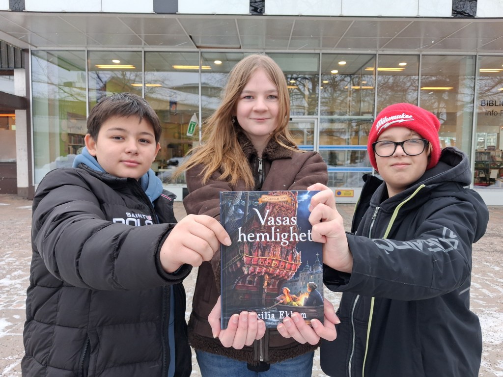 Två pojkar och en flicka håller upp en bok mot kameran.