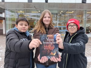 Två pojkar och en flicka håller upp en bok mot kameran.