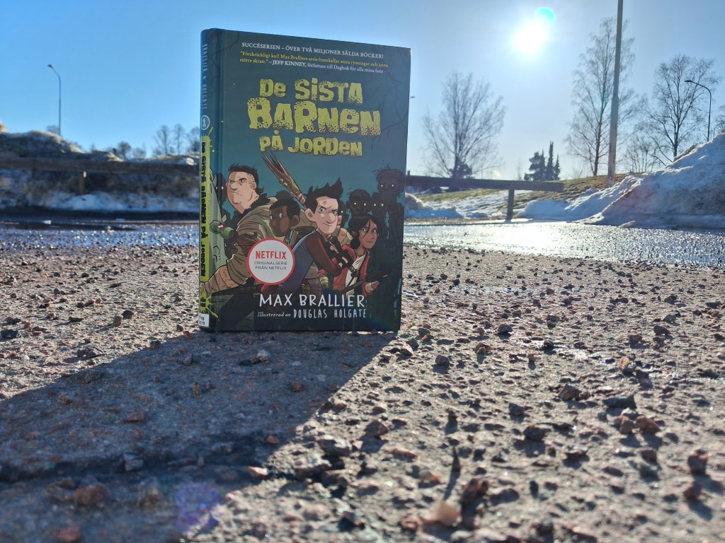 Bok som står uppställd på en grusig asfaltsyta i motljus. I bakgrunden en snödriva som smälter och blå himmel.